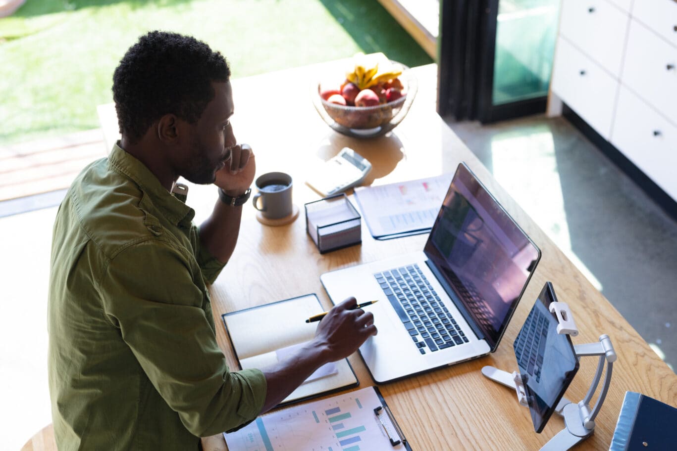 Happy african american man standing at table in kitchen, using laptop and tablet. Spending quality time at home alone.